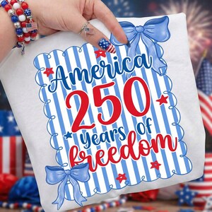 May include: White t-shirt with a patriotic design celebrating 250 years of freedom. The design features the word "America" in blue, the number "250" in red, and the words "years of freedom" in red. Blue bows and red stars decorate the design.