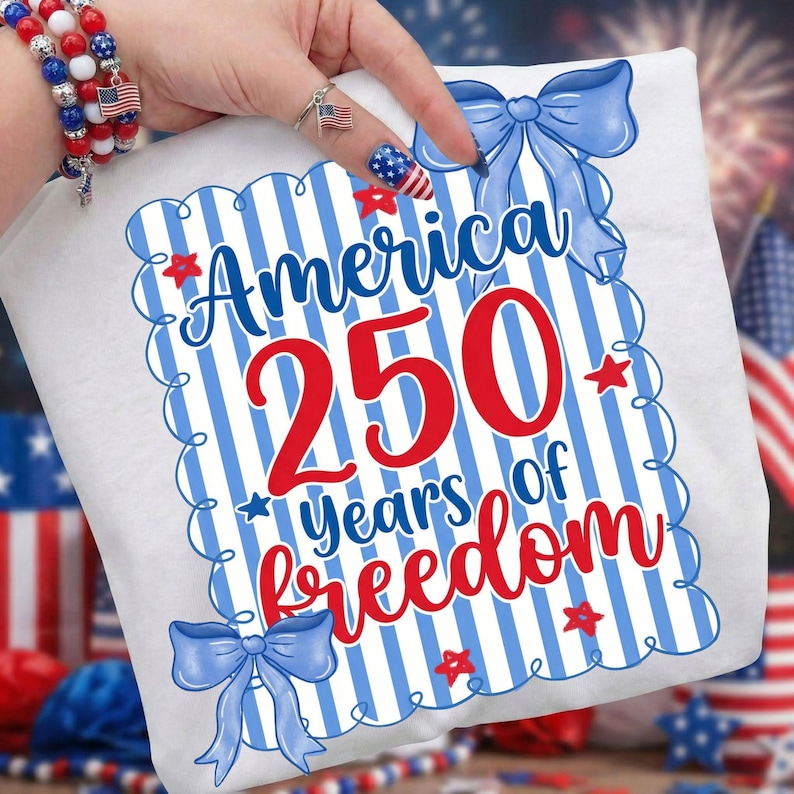 May include: White t-shirt with a patriotic design celebrating 250 years of freedom. The design features the word "America" in blue, the number "250" in red, and the words "years of freedom" in red. Blue bows and red stars decorate the design.