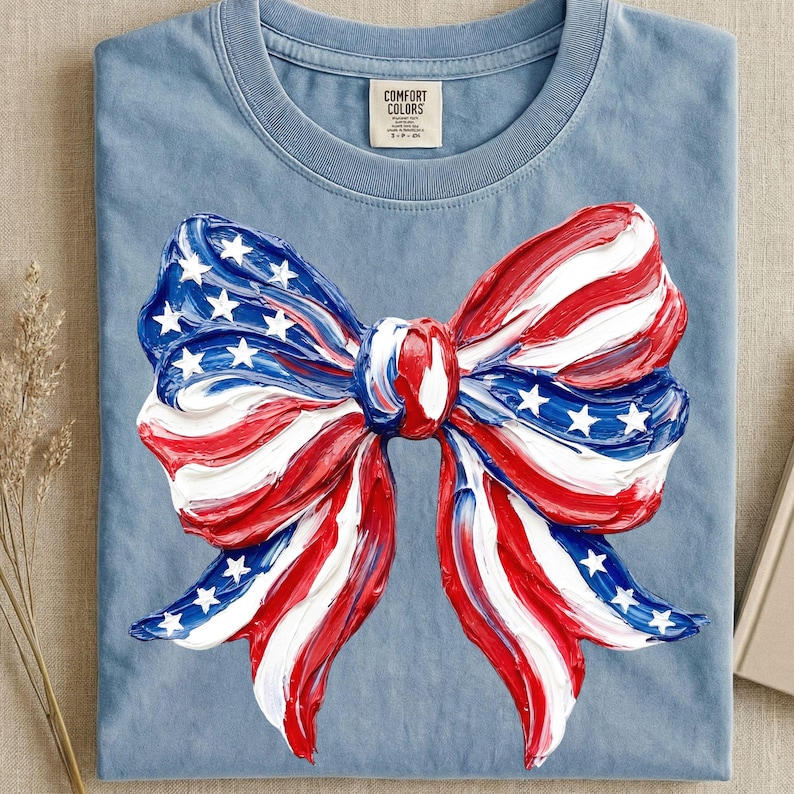 May include: A light blue t-shirt featuring a large bow design in red, white, and blue, resembling the American flag. The bow has white stars on a blue background and red and white stripes. The shirt has a crew neck and a tag that says "Comfort Colors".
