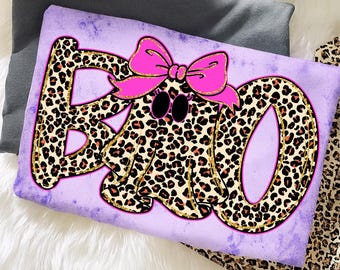 May include: A lavender pillow with a leopard print "BOO" ghost design. The letters and ghost are covered in a leopard print pattern with gold glitter trim. A pink bow is on top of the ghost's head. The background is a white fluffy surface.