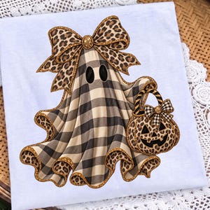 May include: A white t-shirt featuring a graphic of a ghost wearing a large leopard print bow. The ghost has a black and beige buffalo plaid pattern, two black oval eyes, and carries a leopard print pumpkin bucket with a plaid bow.