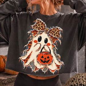 Op de afbeelding: Een antracietgrijs sweatshirt met lange mouwen en een applicatie met gerafelde randen van een spook met een strik met luipaardprint die een oranje pompoenemmer vasthoudt. Het ontwerp bevat stoffen patches met ruiten en strepen.