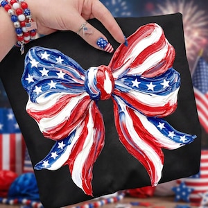 May include: A black pillow featuring a large, textured bow design in red, white, and blue, reminiscent of the American flag. The bow has white stars on a blue background. The pillow is a patriotic decorative item.
