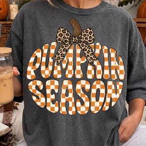 May include: A charcoal gray short-sleeve t-shirt featuring a large graphic of a pumpkin shape filled with an orange and white checkered pattern. The text PUMPKIN SEASON is written in a bold font across the pumpkin, topped with a leopard print bow.