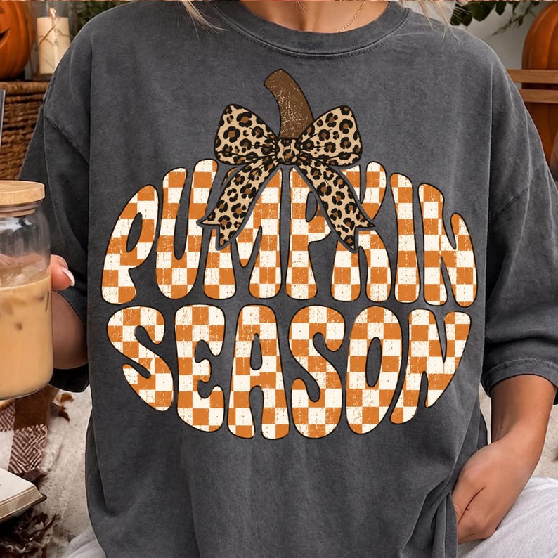 May include: A charcoal gray short-sleeve t-shirt featuring a large graphic of a pumpkin shape filled with an orange and white checkered pattern. The text PUMPKIN SEASON is written in a bold font across the pumpkin, topped with a leopard print bow.