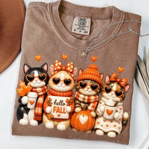 May include: A brown t-shirt featuring a graphic of four cartoon cats wearing heart-shaped sunglasses and autumn-themed accessories. The cats are holding a sign that reads "hello FALL" and various items including a maple leaf, a pumpkin, and mugs.