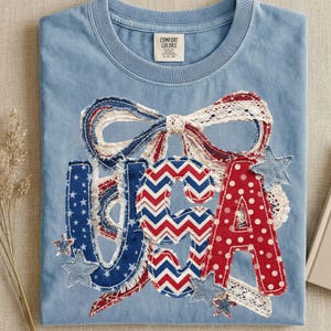 May include: Light blue t-shirt with a patriotic USA design. The letters are in red, white, and blue patterns, with a bow and stars. The shirt is made of a soft material and is perfect for celebrating Independence Day.