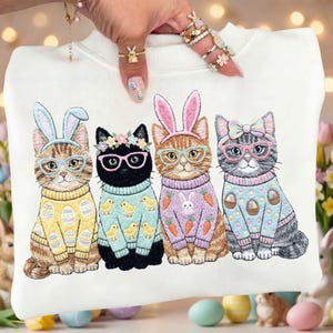 Puede incluir: Sudadera blanca con un diseño bordado de cuatro gatos vestidos para Pascua. Cada gato lleva gafas y un suéter con diseños de Pascua. Los gatos tienen orejas de conejo y coronas florales.