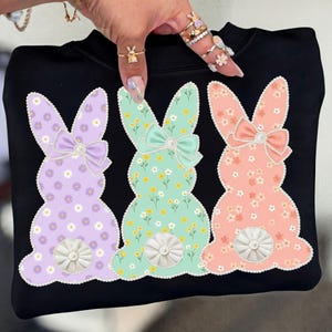 Peut inclure: Sweat-shirt noir orné de trois lapins aux couleurs pastel. Chaque lapin présente un motif floral, un nœud et une queue décorative. Les lapins sont de couleur lavande, vert menthe et pêche.