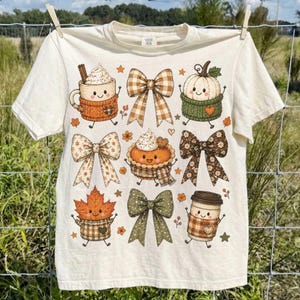 May include: A cream-colored t-shirt featuring a graphic print of various autumn-themed characters including a coffee mug, a pumpkin, a pie, a maple leaf, and a travel cup, all interspersed with several patterned bows and small orange stars.
