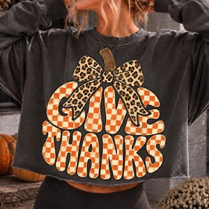 May include: A charcoal gray long-sleeve sweatshirt featuring a large graphic of a pumpkin shape filled with an orange and white checkered pattern. A leopard print bow sits atop the pumpkin, and the text GIVE THANKS is written in orange and white.