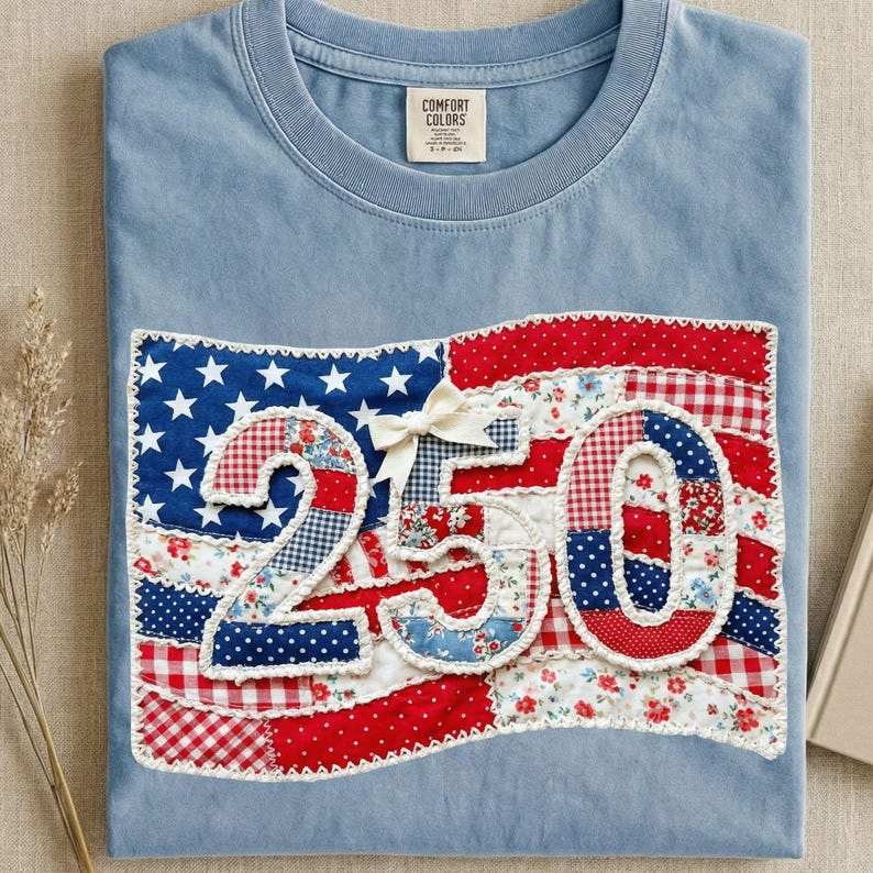 May include: Light blue t-shirt with a patriotic design. The design features a patchwork American flag with the number "250" in the center, made of various patterned fabrics. A white bow is attached to the flag.