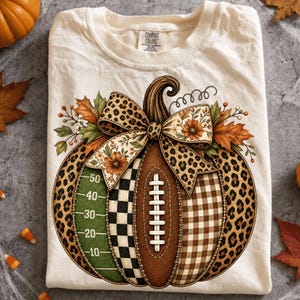 Peut inclure: Un t-shirt couleur crème avec une étiquette Comfort Colors présentant un motif de citrouille décoré d'imprimé léopard, d'un ballon de football, d'un terrain de football vert avec des lignes, de carreaux noirs et blancs et de vichy marron.