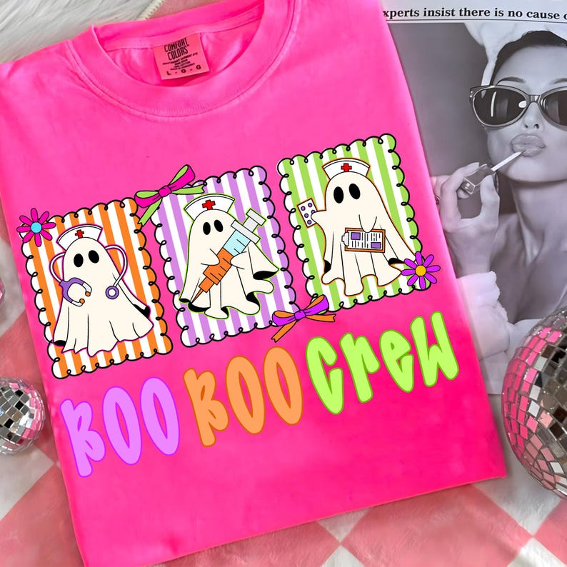 May include: Bright pink t-shirt with a graphic of three cartoon ghosts dressed as nurses, each with medical tools. The text "BOO BOO CREW" is in colorful letters below the ghosts. The shirt has a crew neck and short sleeves.