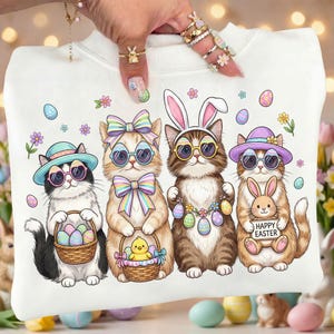 May include: White sweatshirt featuring a colorful Easter-themed graphic. The design showcases four cartoon cats wearing sunglasses and Easter accessories, holding baskets of eggs, a chick, and a sign that says "Happy Easter".