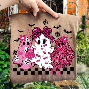 May include: A brown tote bag featuring three ghost characters with pink and black accents. The ghosts have various patterns, including leopard print, cherries, and hearts. Bats, pumpkins, and spiderwebs complete the Halloween theme.