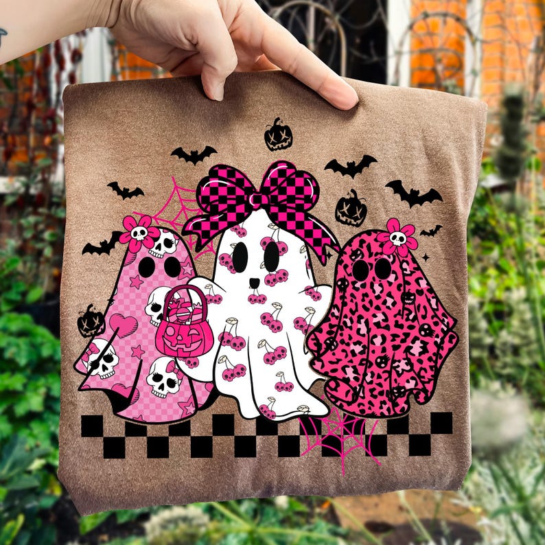 May include: A brown tote bag featuring three ghost characters with pink and black accents. The ghosts have various patterns, including leopard print, cherries, and hearts. Bats, pumpkins, and spiderwebs complete the Halloween theme.