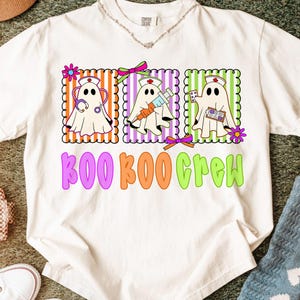 May include: White t-shirt with a colorful graphic of three ghost characters dressed as nurses, each holding medical tools. The text "BOO BOO CREW" is printed below the ghosts in purple, orange, and green.