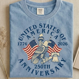 May include: Light blue t-shirt with a graphic commemorating the 250th anniversary of the United States of America. The design includes an eagle, flags, the Statue of Liberty, and the text "United States of America Anniversary 1776-2026".