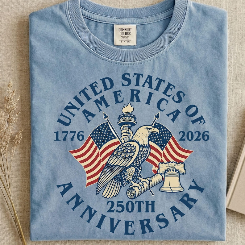 May include: Light blue t-shirt with a graphic commemorating the 250th anniversary of the United States of America. The design includes an eagle, flags, the Statue of Liberty, and the text "United States of America Anniversary 1776-2026".