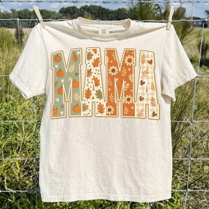 May include: A cream-colored short-sleeve t-shirt featuring the word MAMA printed in large block letters across the chest. Each letter contains a different autumn-themed pattern, including orange pumpkins, fall leaves, sunflowers, and a beige plaid.