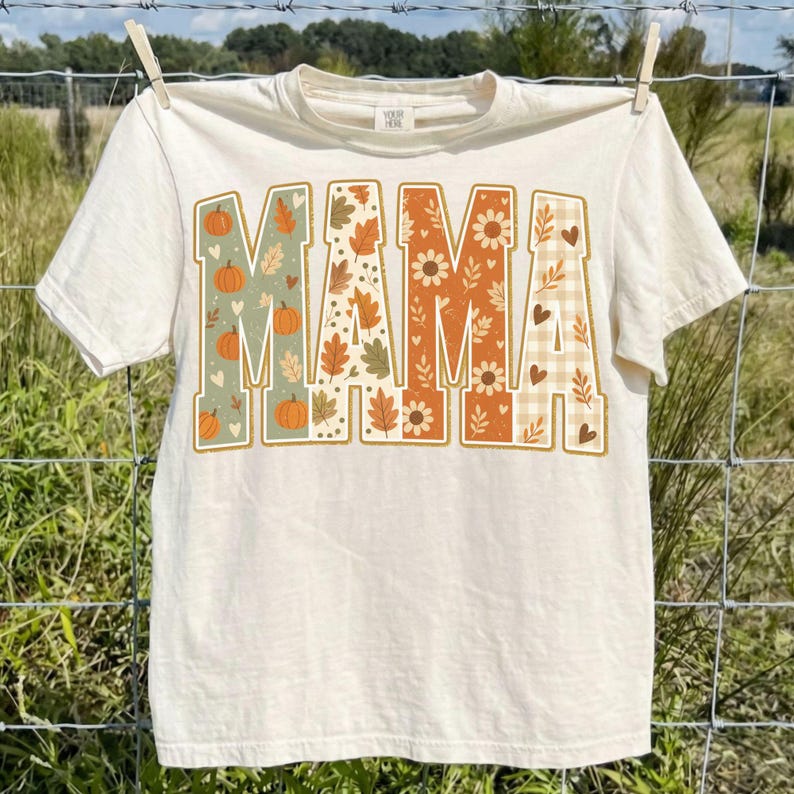 May include: A cream-colored short-sleeve t-shirt featuring the word MAMA printed in large block letters across the chest. Each letter contains a different autumn-themed pattern, including orange pumpkins, fall leaves, sunflowers, and a beige plaid.