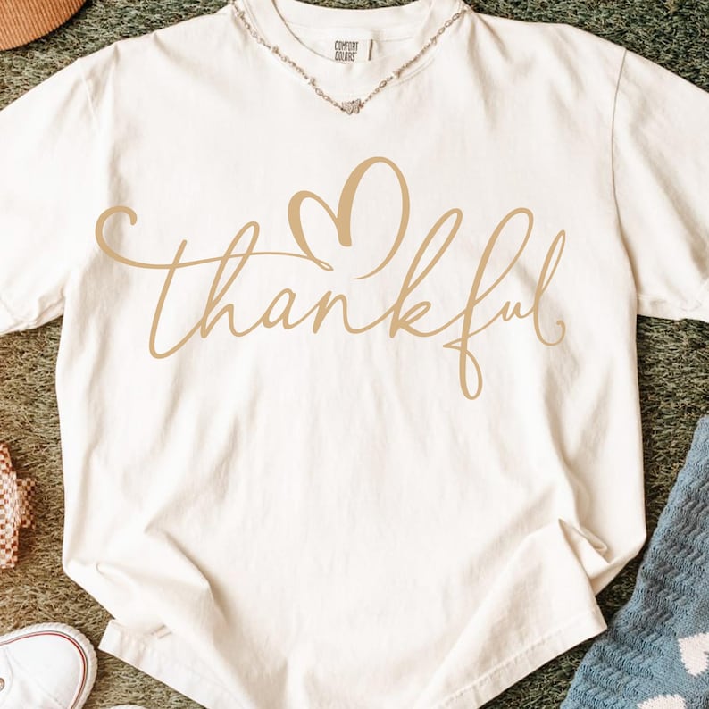 May include: Cream-colored t-shirt with the word "thankful" in a cursive font, featuring a heart-shaped design above the word. The shirt is made of a soft material and is suitable for casual wear.
