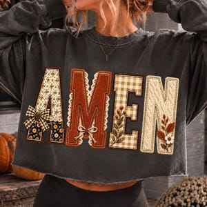 May include: A charcoal gray long-sleeve sweatshirt featuring the word AMEN in large, textured applique letters. Each letter has a unique pattern, including floral prints, corduroy, gingham, and cable knit, accented with small bows and floral sprigs.