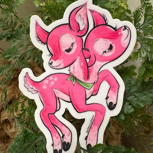 May include: A sticker featuring a pink two-headed deer illustration with a green bow. The deer has closed eyes and white spots. The sticker is surrounded by green leaves.