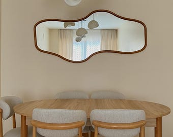 May include: A dining room with an oval wooden table and six chairs with light grey upholstery. Above the table is a large, uniquely shaped mirror with a brown frame, and three white pendant lights.