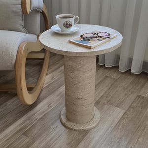 May include: A round beige travertine side table with a cylindrical pedestal base. On the tabletop sits a white ceramic teacup with a floral pattern on a matching saucer, a small rectangular book, and a pair of brown tortoiseshell reading glasses.