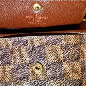 May include: Close-up of a brown and tan checkered wallet with a gold snap closure. The top flap is open, revealing the interior and the embossed text "LOUIS VUITTON PARIS made in France".