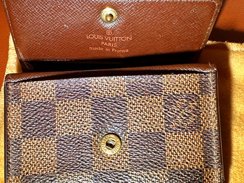 May include: Close-up of a brown and tan checkered wallet with a gold snap closure. The top flap is open, revealing the interior and the embossed text "LOUIS VUITTON PARIS made in France".