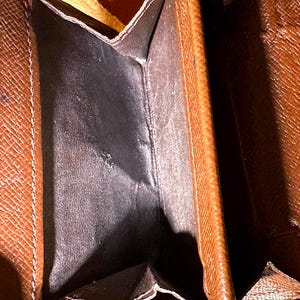 May include: Open brown wallet with a dark interior. The wallet has a textured exterior and visible stitching along the edges. The interior appears to be made of a smooth, dark material.