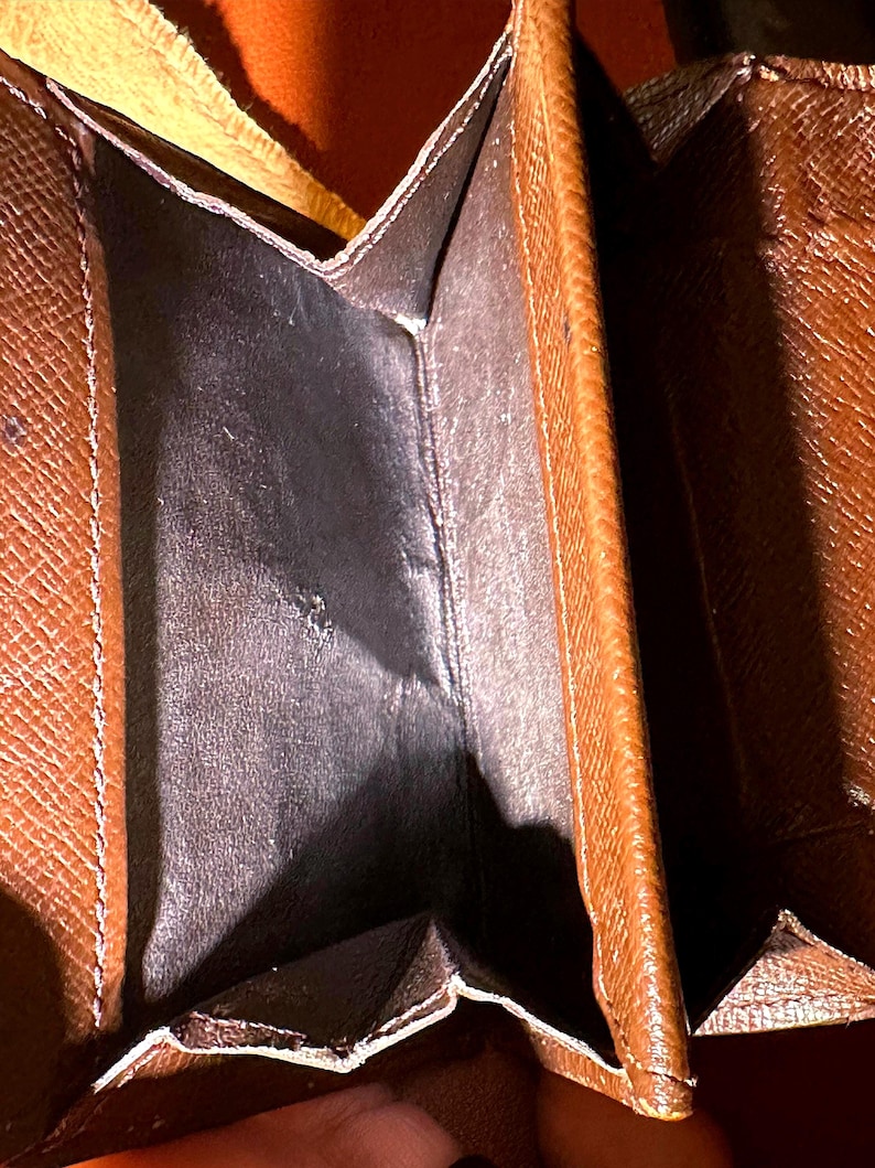 May include: Open brown wallet with a dark interior. The wallet has a textured exterior and visible stitching along the edges. The interior appears to be made of a smooth, dark material.