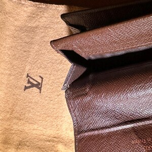 May include: Brown leather wallet with a monogram pattern, partially open, revealing the interior. A tan fabric pouch with a logo is visible in the background. The wallet appears to be made of high-quality materials.