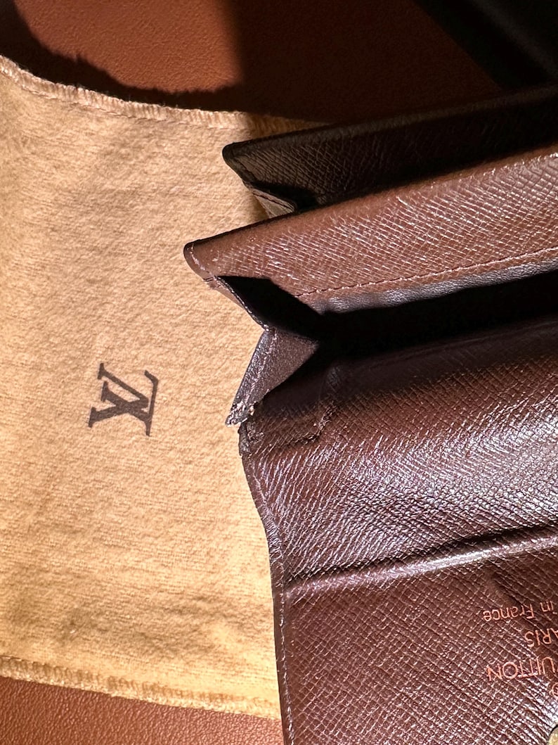 May include: Brown leather wallet with a monogram pattern, partially open, revealing the interior. A tan fabric pouch with a logo is visible in the background. The wallet appears to be made of high-quality materials.