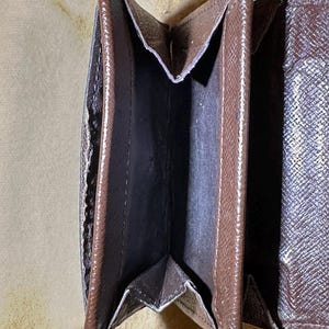 May include: Open brown wallet with multiple compartments. The wallet has a textured exterior and a dark interior. The wallet is open, revealing the interior pockets and the lining. The wallet is on a light brown surface.