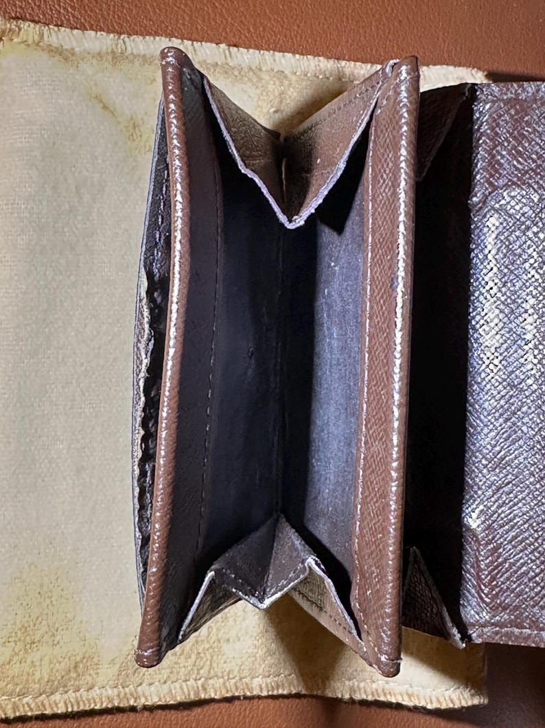 May include: Open brown wallet with multiple compartments. The wallet has a textured exterior and a dark interior. The wallet is open, revealing the interior pockets and the lining. The wallet is on a light brown surface.