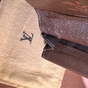 May include: Brown leather wallet with a textured pattern, open to reveal a tan fabric pouch with the Louis Vuitton logo. The wallet has a gold-colored clasp and the words "LOUIS VUITTON PARIS made in France" are visible.