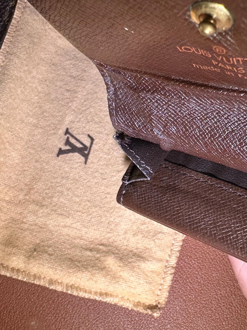 May include: Brown leather wallet with a textured pattern, open to reveal a tan fabric pouch with the Louis Vuitton logo. The wallet has a gold-colored clasp and the words "LOUIS VUITTON PARIS made in France" are visible.
