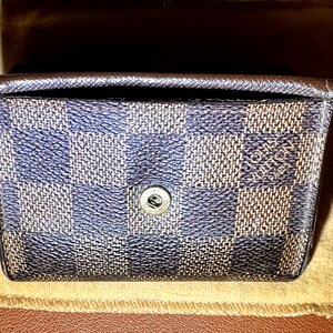 May include: A small, rectangular wallet with a checkered pattern in shades of blue and brown. The wallet has a snap closure and the brand name "LOUIS VUITTON PARIS" is visible. The wallet is resting on a tan cloth.