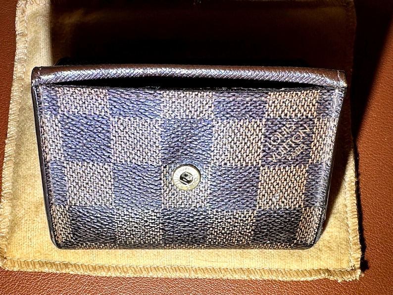 May include: A small, rectangular wallet with a checkered pattern in shades of blue and brown. The wallet has a snap closure and the brand name "LOUIS VUITTON PARIS" is visible. The wallet is resting on a tan cloth.