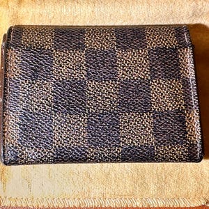 May include: A small, square wallet with a checkered pattern in shades of brown and navy blue. The wallet is closed and appears to be made of a textured material. It rests on a light tan cloth.