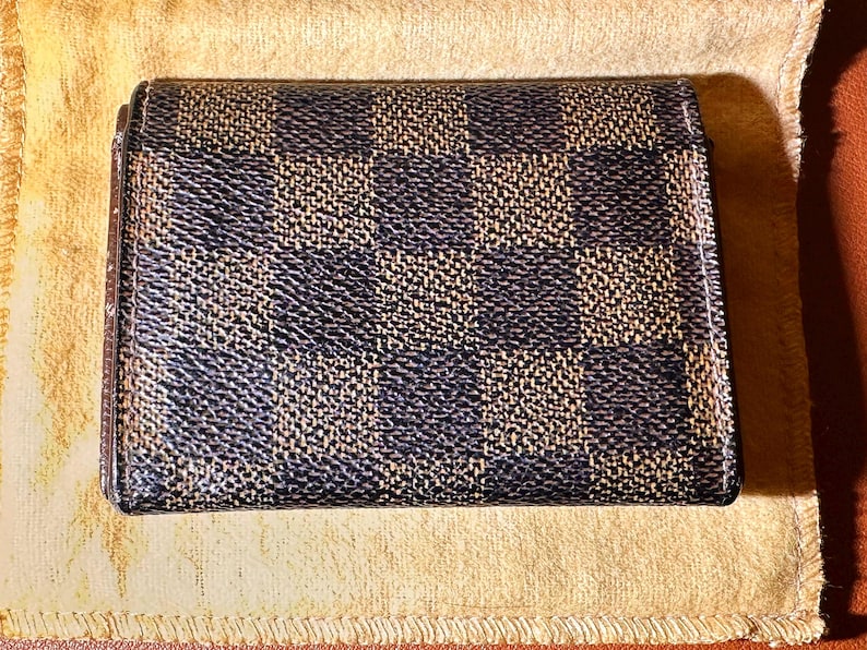 May include: A small, square wallet with a checkered pattern in shades of brown and navy blue. The wallet is closed and appears to be made of a textured material. It rests on a light tan cloth.