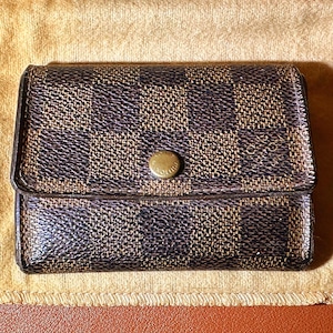 May include: A brown and black checkered wallet with a gold button clasp. The wallet is rectangular and sits on a tan cloth. The pattern is a repeating square design.