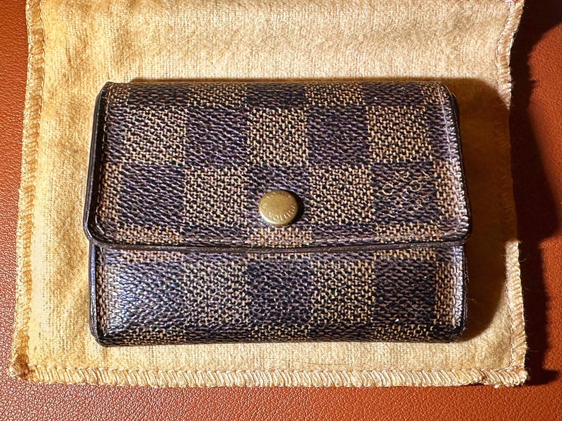 May include: A brown and black checkered wallet with a gold button clasp. The wallet is rectangular and sits on a tan cloth. The pattern is a repeating square design.