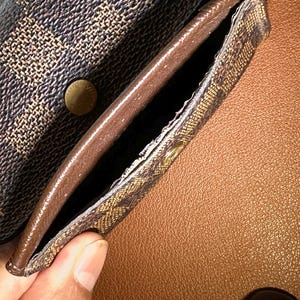 May include: Close-up of a brown and tan patterned wallet with a snap closure. The interior is a solid brown color. The wallet is open, revealing a pocket. The background is a textured brown surface.