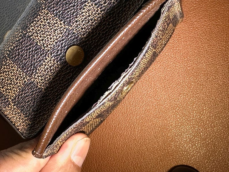 May include: Close-up of a brown and tan patterned wallet with a snap closure. The interior is a solid brown color. The wallet is open, revealing a pocket. The background is a textured brown surface.