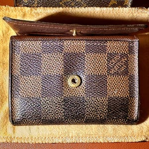 May include: A brown and black checkered wallet with a gold button clasp. The wallet is open, revealing a brown interior. The wallet is resting on a tan cloth.